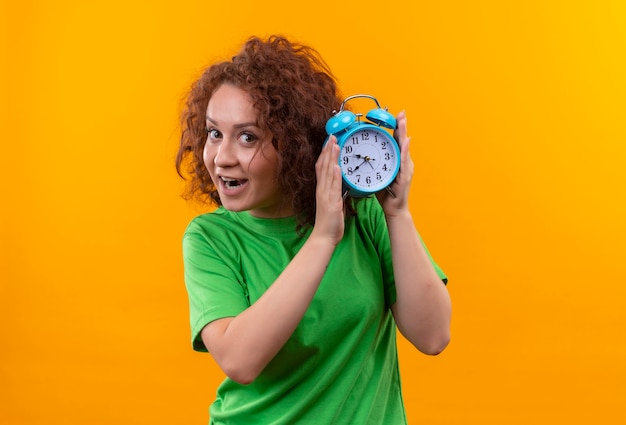 Mulher jovem com cabelo curto e encaracolado com uma camiseta verde segurando um despertador feliz e surpresa em pé sobre uma parede laranja