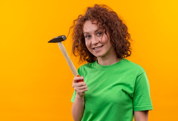 Mulher jovem com cabelo curto e encaracolado com uma camiseta verde segurando um martelo e sorrindo alegremente em pé