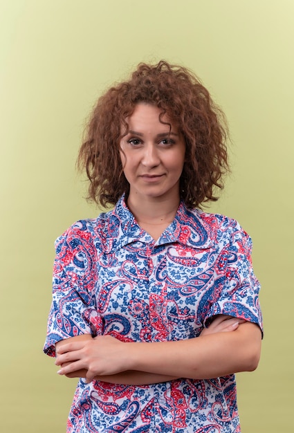 Foto grátis mulher jovem com cabelo curto e encaracolado em uma camisa colorida em pé com os braços cruzados no peito e olhando com uma cara séria sobre a parede verde