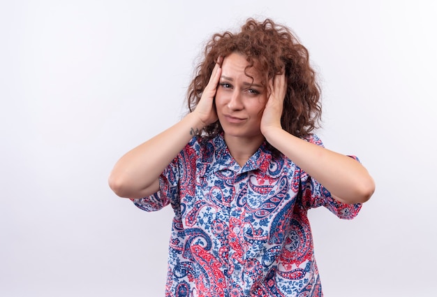 Foto grátis mulher jovem com cabelo curto e encaracolado em uma camisa colorida estressada e preocupada tocando a cabeça com as mãos em pé sobre uma parede branca