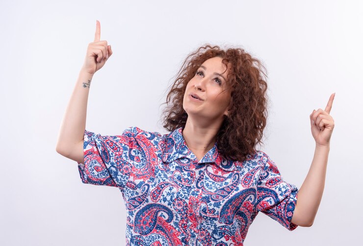 Foto grátis mulher jovem com cabelo curto e encaracolado em uma camisa colorida feliz e positiva olhando para cima e apontando para cima com os dedos em pé sobre a parede branca