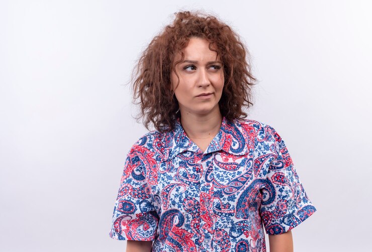 Foto grátis mulher jovem com cabelo curto e encaracolado em uma camisa colorida olhando para o lado com expressão cética em pé sobre uma parede branca