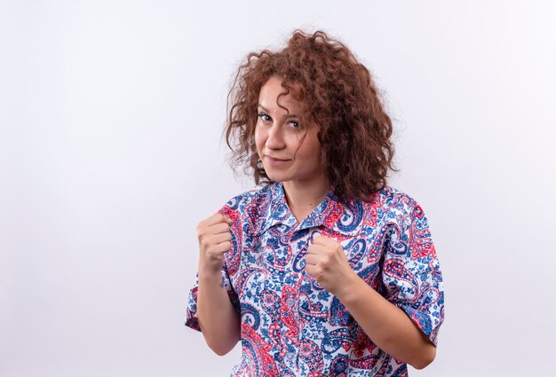 Mulher jovem com cabelo curto e encaracolado em uma camisa colorida, parecendo confiante com os punhos cerrados, como um boxeador em pé sobre uma parede branca