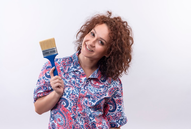 Foto grátis mulher jovem com cabelo curto e encaracolado em uma camisa colorida segurando um pincel e sorrindo alegremente em pé sobre uma parede branca