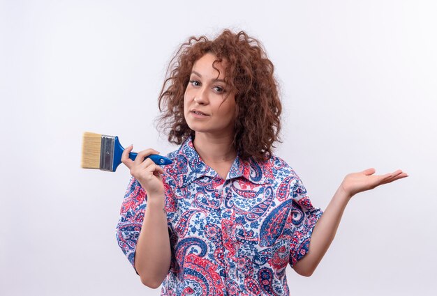 Mulher jovem com cabelo curto e encaracolado em uma camisa colorida segurando um pincel, sorrindo, apresentando-se com o braço da mão em pé sobre uma parede branca