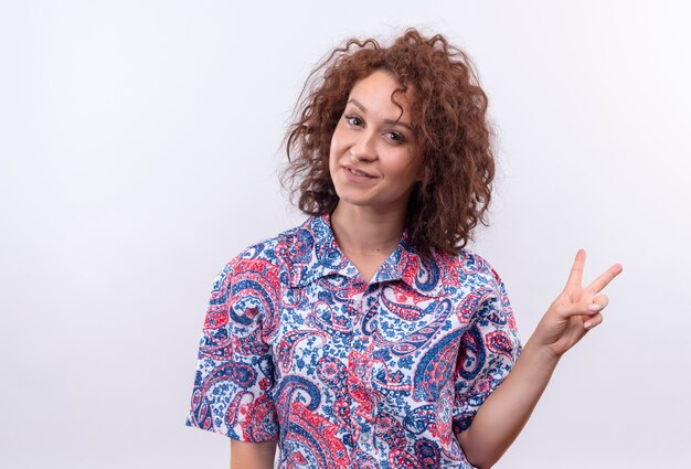 Foto grátis mulher jovem com cabelo curto e encaracolado em uma camisa colorida sorrindo e mostrando o sinal da vitória sobre uma parede branca