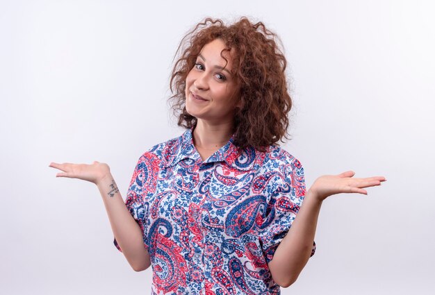 Mulher jovem com cabelo curto e encaracolado em uma camisa colorida sorrindo, olhando se apresentando com os dois braços, oh, mãos em pé sobre uma parede branca