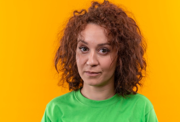 Mulher jovem com cabelo curto e encaracolado em uma camiseta verde com cara séria em pé