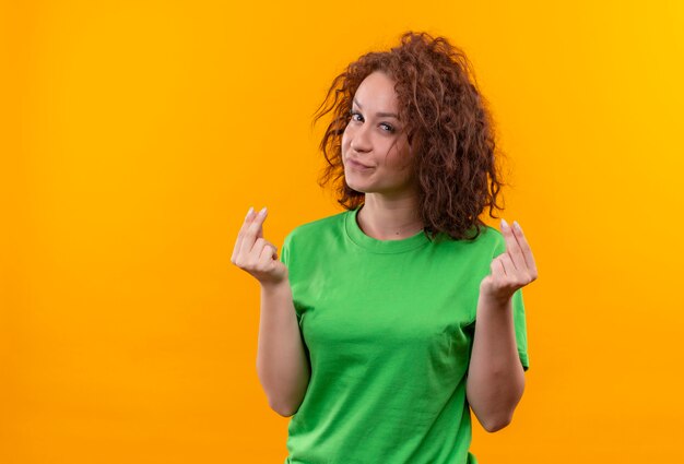 Mulher jovem com cabelo curto e encaracolado em uma camiseta verde esfregando os dedos fazendo gesto de dinheiro pedindo dinheiro em pé sobre uma parede laranja