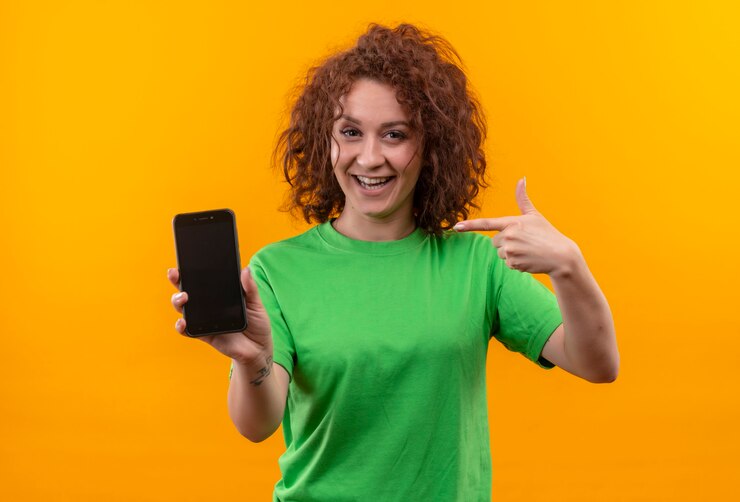 Foto grátis mulher jovem com cabelo curto e encaracolado em uma camiseta verde mostrando smartphone apontando com o dedo para ele e sorrindo alegremente em pé sobre uma parede laranja