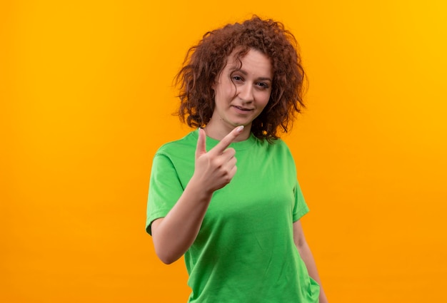 Mulher jovem com cabelo curto e encaracolado em uma camiseta verde mostrando um dedo sorrindo em pé sobre uma parede laranja