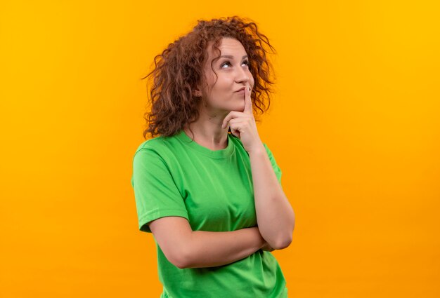 Mulher jovem com cabelo curto e encaracolado em uma camiseta verde olhando com expressão pensativa no rosto pensando em pé sobre a parede laranja