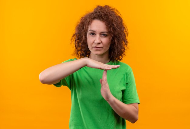 Mulher jovem com cabelo curto e encaracolado em uma camiseta verde parecendo cansada fazendo gesto de pausa