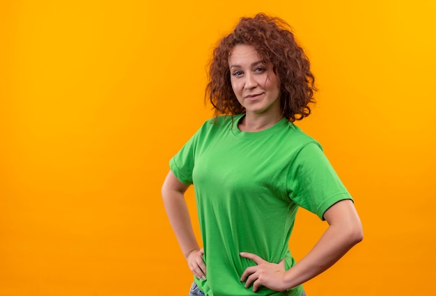 Mulher jovem com cabelo curto e encaracolado em uma camiseta verde parecendo confiante com um sorriso no rosto em pé