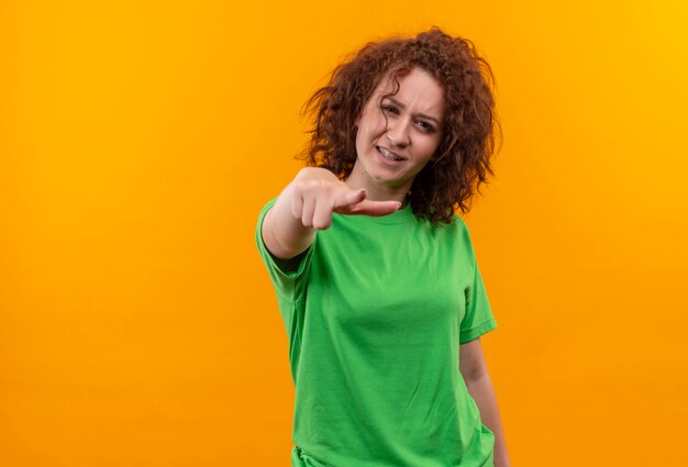 Mulher jovem com cabelo curto e encaracolado em uma camiseta verde parecendo descontente apontando com o dedo indicador na frente