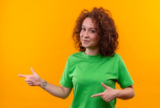 Mulher jovem com cabelo curto e encaracolado em uma camiseta verde sorrindo alegremente apontando com os dedos para o lado em pé sobre uma parede laranja