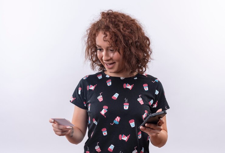 Foto grátis mulher jovem com cabelo curto e encaracolado segurando um cartão de crédito e um smartphone, parecendo surpresa e feliz em pé sobre uma parede branca