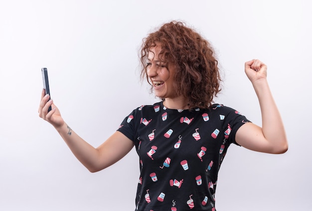 Mulher jovem com cabelo curto e encaracolado segurando um smartphone com o punho cerrado, feliz e animada, comemorando seu sucesso em pé sobre uma parede branca