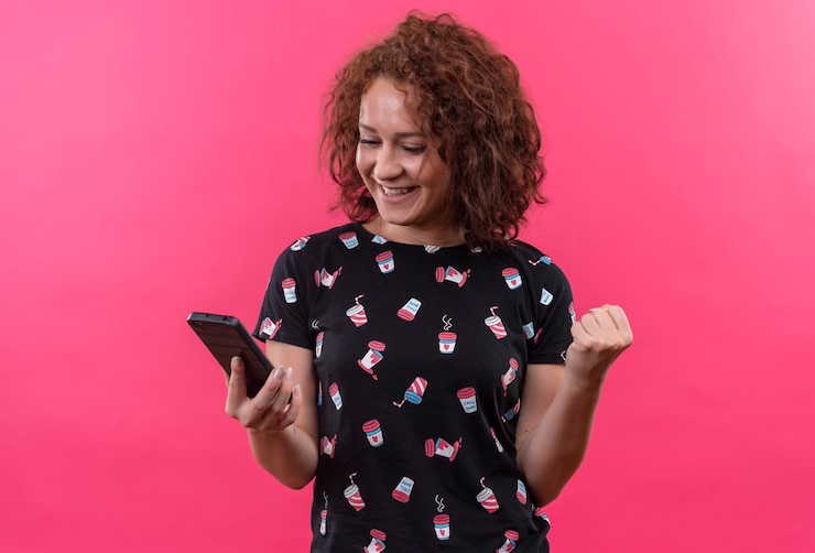 Foto grátis mulher jovem com cabelo curto e encaracolado segurando um smartphone olhando para a tela feliz e animada com o punho cerrado em pé sobre a parede rosa