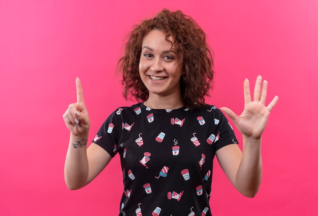 Mulher jovem com cabelo curto e encaracolado sorrindo aparecendo e apontando para cima com os dedos número seis em pé sobre a parede rosa