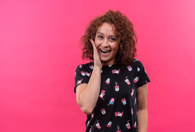 Foto grátis mulher jovem com cabelo curto e encaracolado surpresa e maravilhada, sorrindo amplamente, tocando a bochecha com a mão em pé sobre a parede rosa