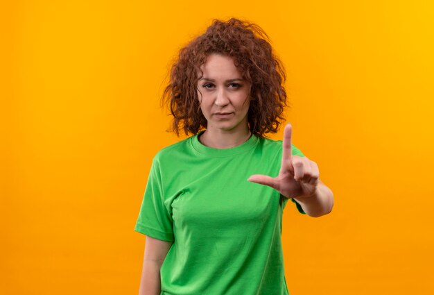 Mulher jovem com cabelo curto e encaracolado usando uma camiseta verde mostrando um aviso com o dedo indicador e uma cara séria em pé sobre uma parede laranja