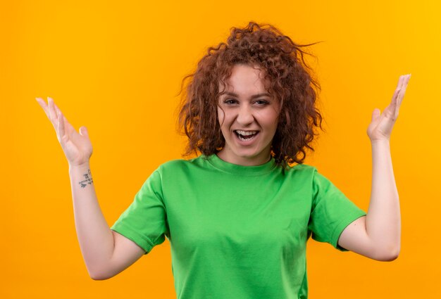 Mulher jovem com cabelo curto e encaracolado usando uma camiseta verde parecendo surpresa e empolgada com os braços levantados em pé