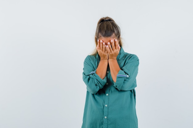 Mulher jovem com camisa azul segurando a mão no rosto e parecendo triste