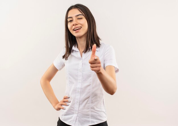 Mulher jovem com camisa branca apontando com o dedo para a frente sorrindo e piscando em pé sobre uma parede branca