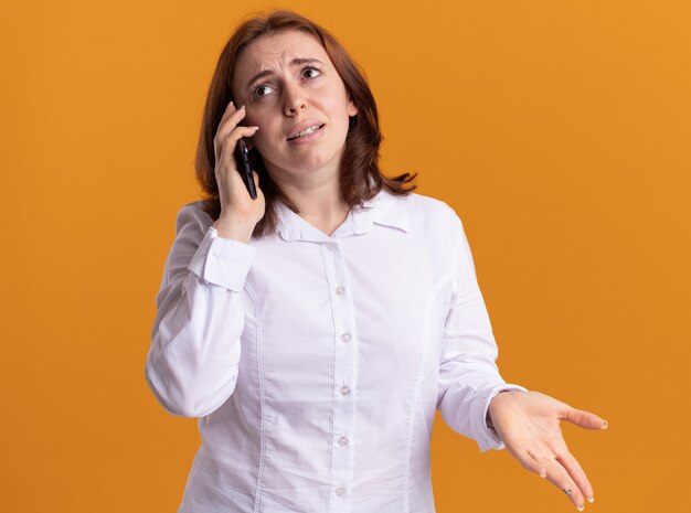 Mulher jovem com camisa branca falando no celular e ficando confusa em pé sobre uma parede laranja