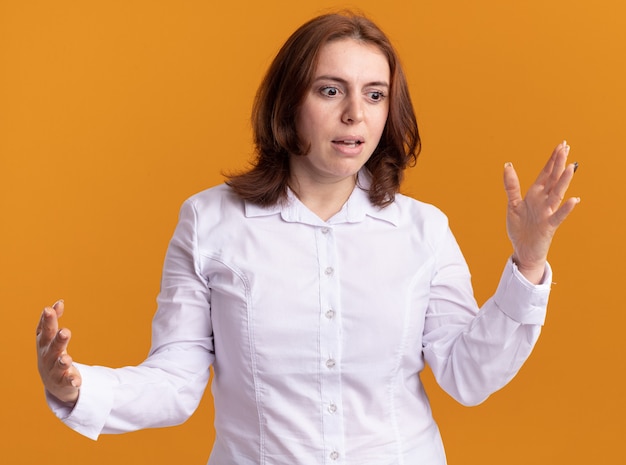 Mulher jovem com camisa branca olhando de lado, confusa, gesticulando com as mãos em pé sobre a parede laranja