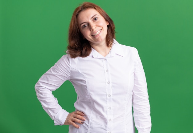 Mulher jovem com camisa branca olhando para a frente feliz e sorridente confiante em pé sobre a parede verde
