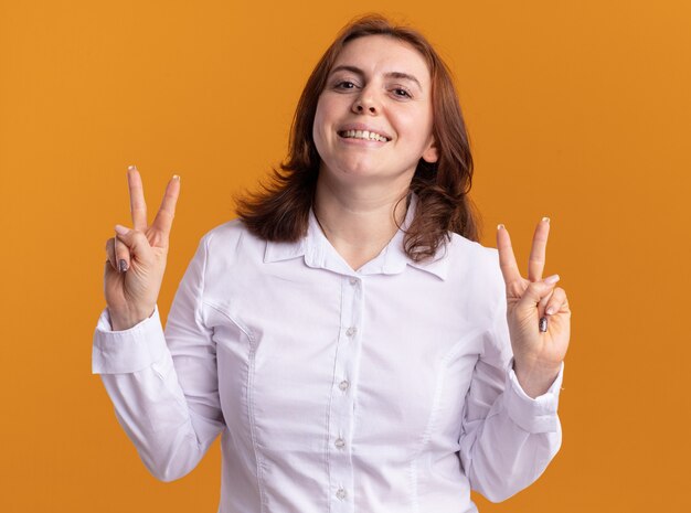 Mulher jovem com camisa branca olhando para a frente feliz e sorrindo positivamente mostrando o sinal-V em pé sobre a parede laranja