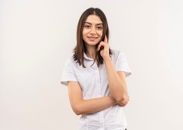 Mulher jovem com camisa branca olhando para a frente sorrindo confiante em pé sobre uma parede branca