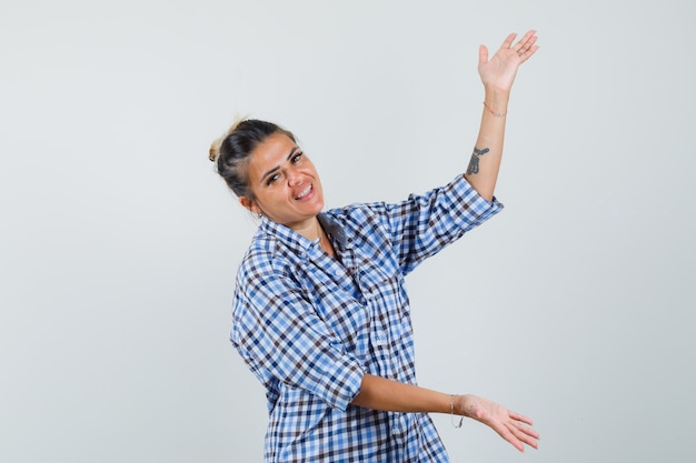 Mulher jovem com camisa quadriculada, espalhando as palmas das mãos de lado enquanto mostra algo e parece feliz.