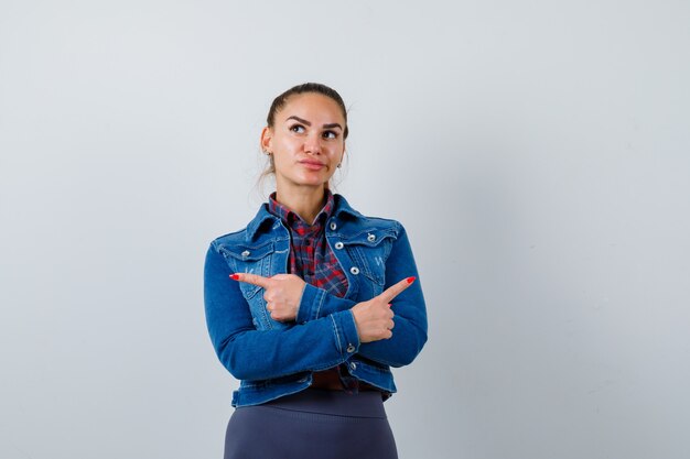 Mulher jovem com camisa quadriculada, jaqueta jeans apontando para o lado com os braços cruzados e olhando pensativa, vista frontal.