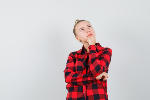 Mulher jovem com camisa xadrez apoiando o queixo no punho levantado e parecendo pensativa