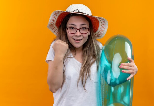 Mulher jovem com camiseta branca e chapéu de verão segurando um anel inflável e cerrando o punho, parecendo animada e feliz em pé sobre a parede laranja