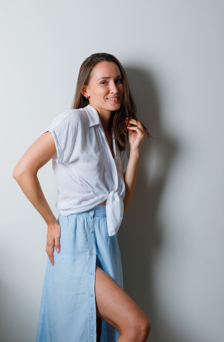 Foto grátis mulher jovem com camiseta branca e saia azul claro segurando a mão no quadril enquanto ajeita o cabelo e parece charmosa