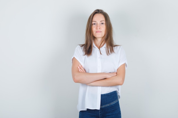 Mulher jovem com camiseta branca, jeans em pé com os braços cruzados e parecendo confiante