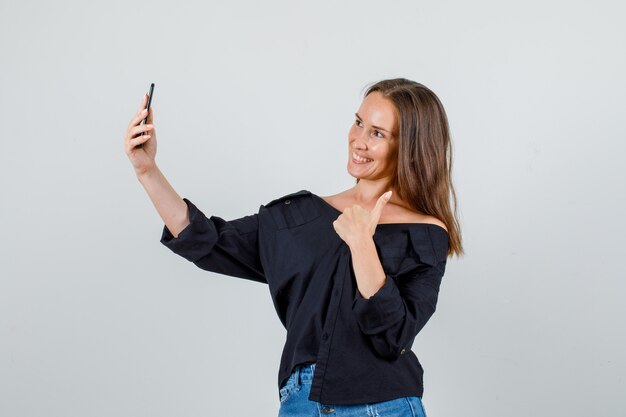 Mulher jovem com camiseta e shorts aparecendo com o polegar enquanto tira uma selfie e parece alegre