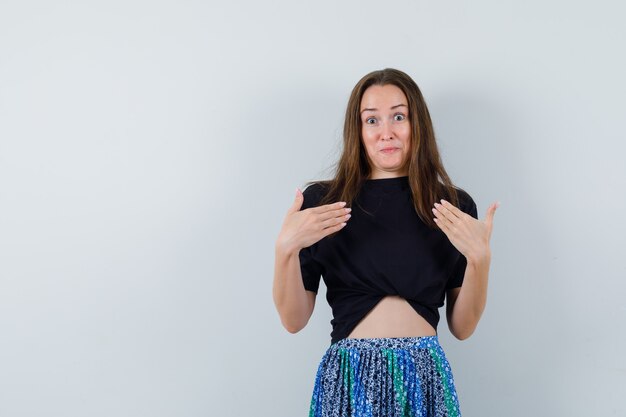 Mulher jovem com camiseta preta e saia azul apontando para si mesma com as duas mãos e parecendo surpresa