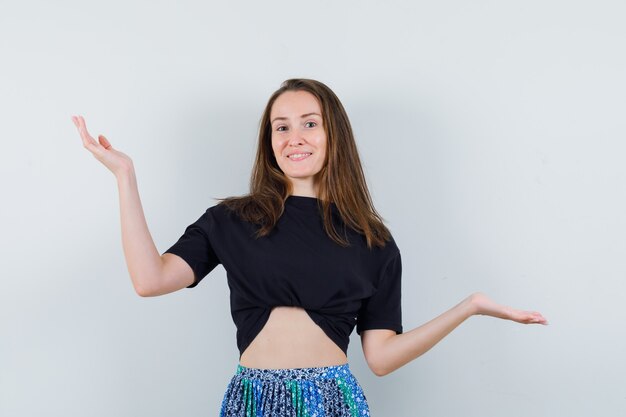 Mulher jovem com camiseta preta e saia azul, mostrando um gesto impotente e parecendo feliz