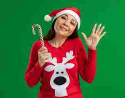 Foto grátis mulher jovem com chapéu de papai noel de natal e suéter vermelho segurando um bastão de doces, olhando para a câmera, feliz e alegre, acenando com a mão em pé sobre um fundo verde