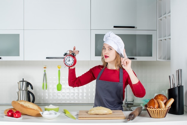 Mulher jovem com chapéu e avental de cozinheira segurando um despertador vermelho e se divertindo com uma ideia na cozinha.