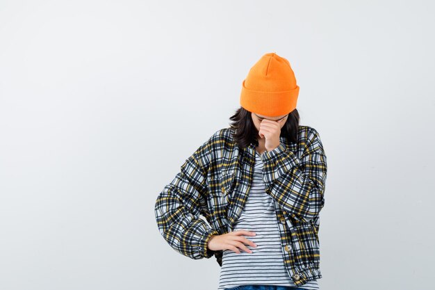 Mulher jovem com chapéu laranja e camisa quadriculada, inclinando a cabeça sobre a mão e parecendo chateada