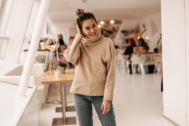 Mulher jovem com coque e vestida com suéter bege e calça jeans escura posando em um café