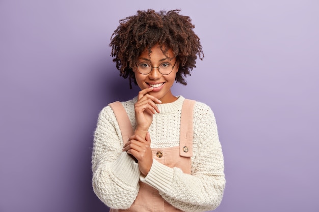 Mulher jovem com corte de cabelo afro, vestindo suéter branco e macacão rosa