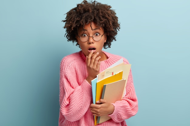 Mulher jovem com corte de cabelo afro, vestindo um suéter rosa e segurando livros didáticos