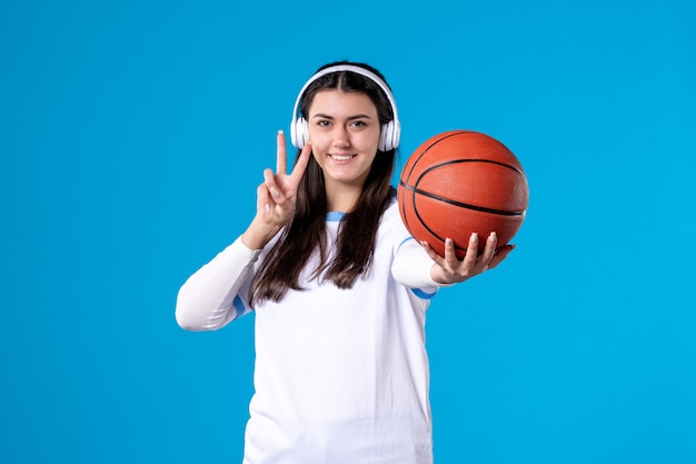 Mulher jovem com fones de ouvido segurando uma bola de basquete na parede azul de frente